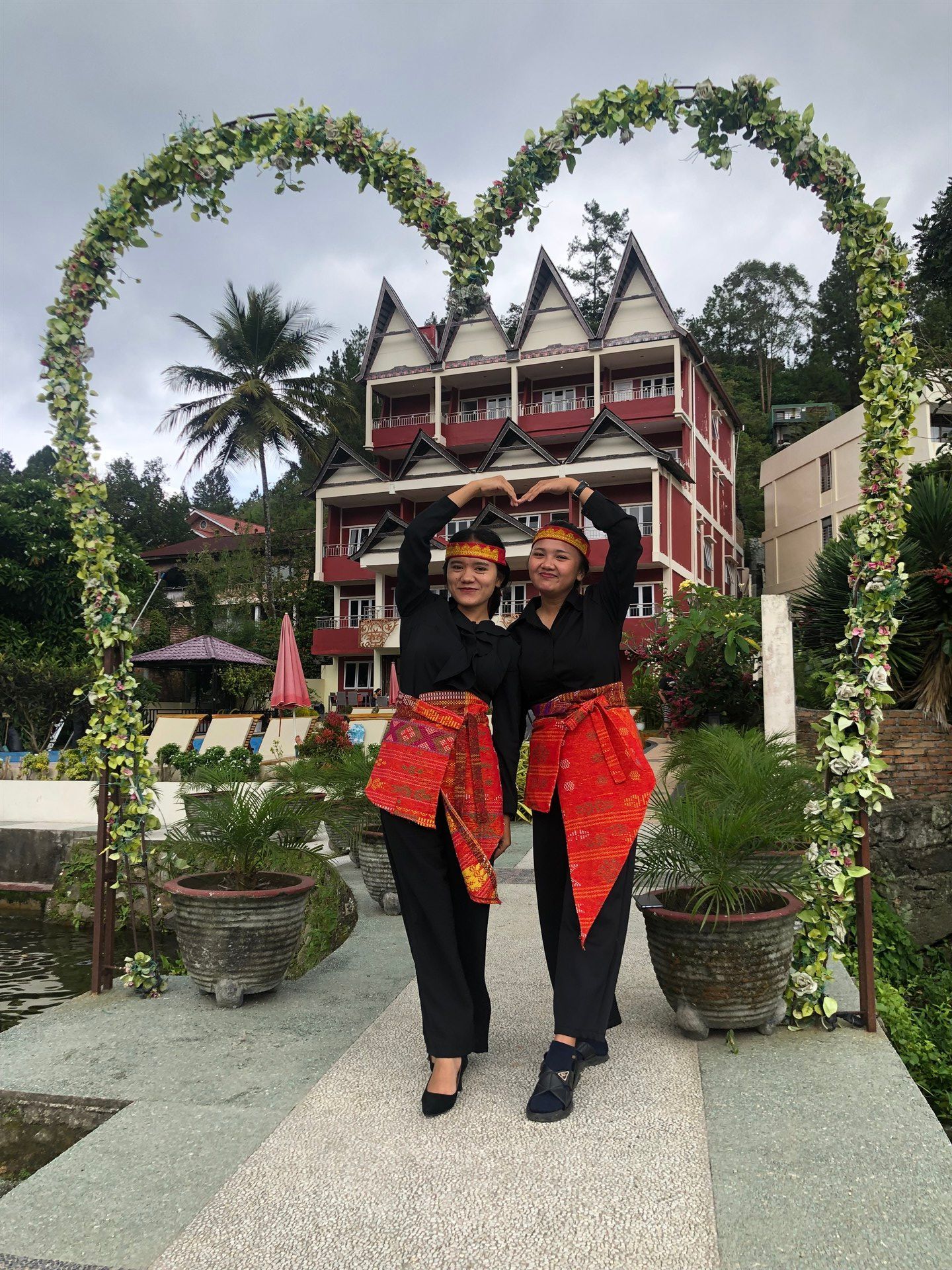 Two women are standing under a heart shaped arch in front of a building.