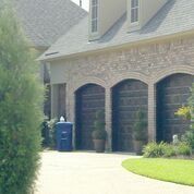 A brick house with three garage doors and a blue trash can in front of it.