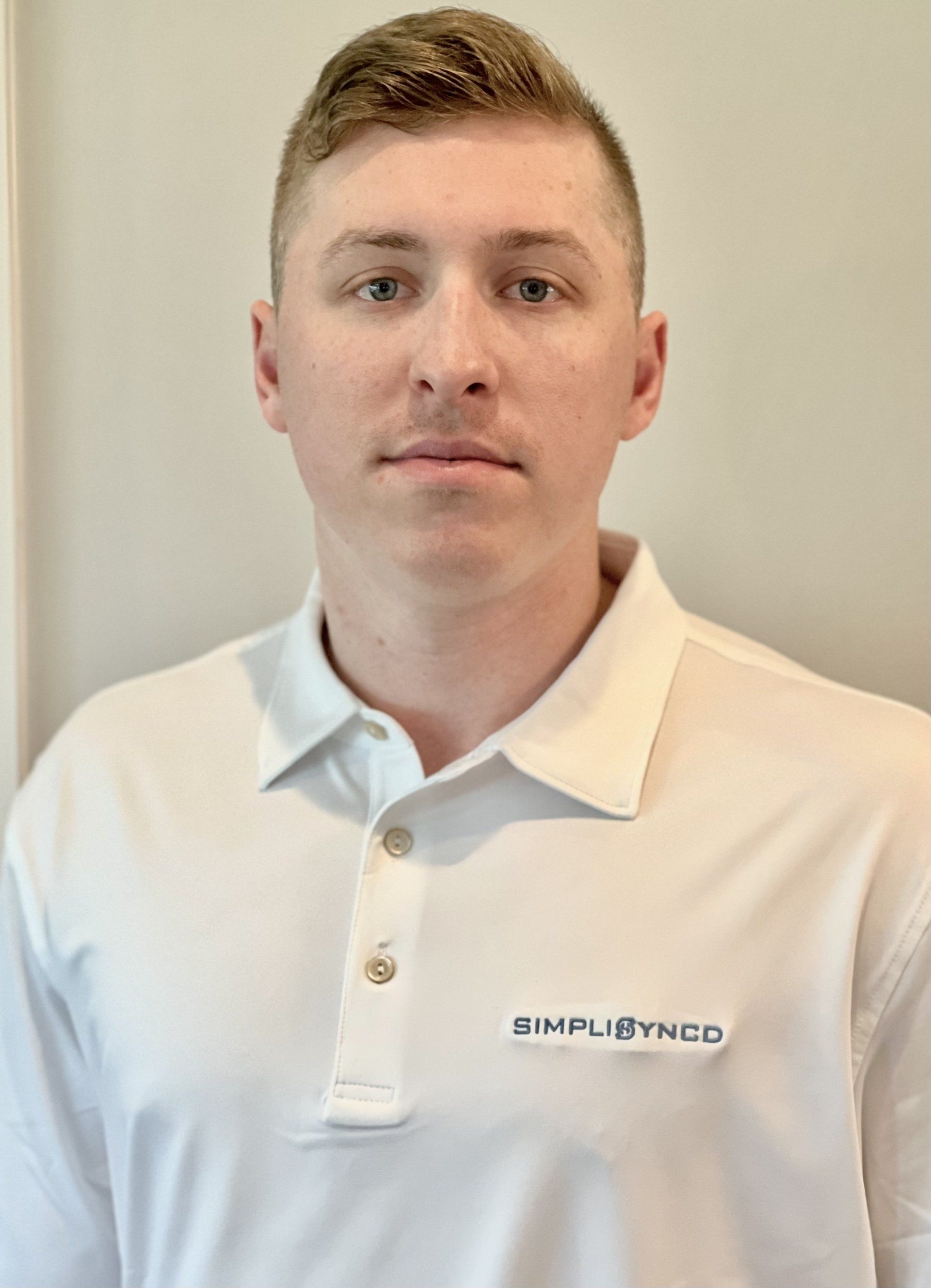 A man wearing a white polo shirt is standing in front of a white wall.