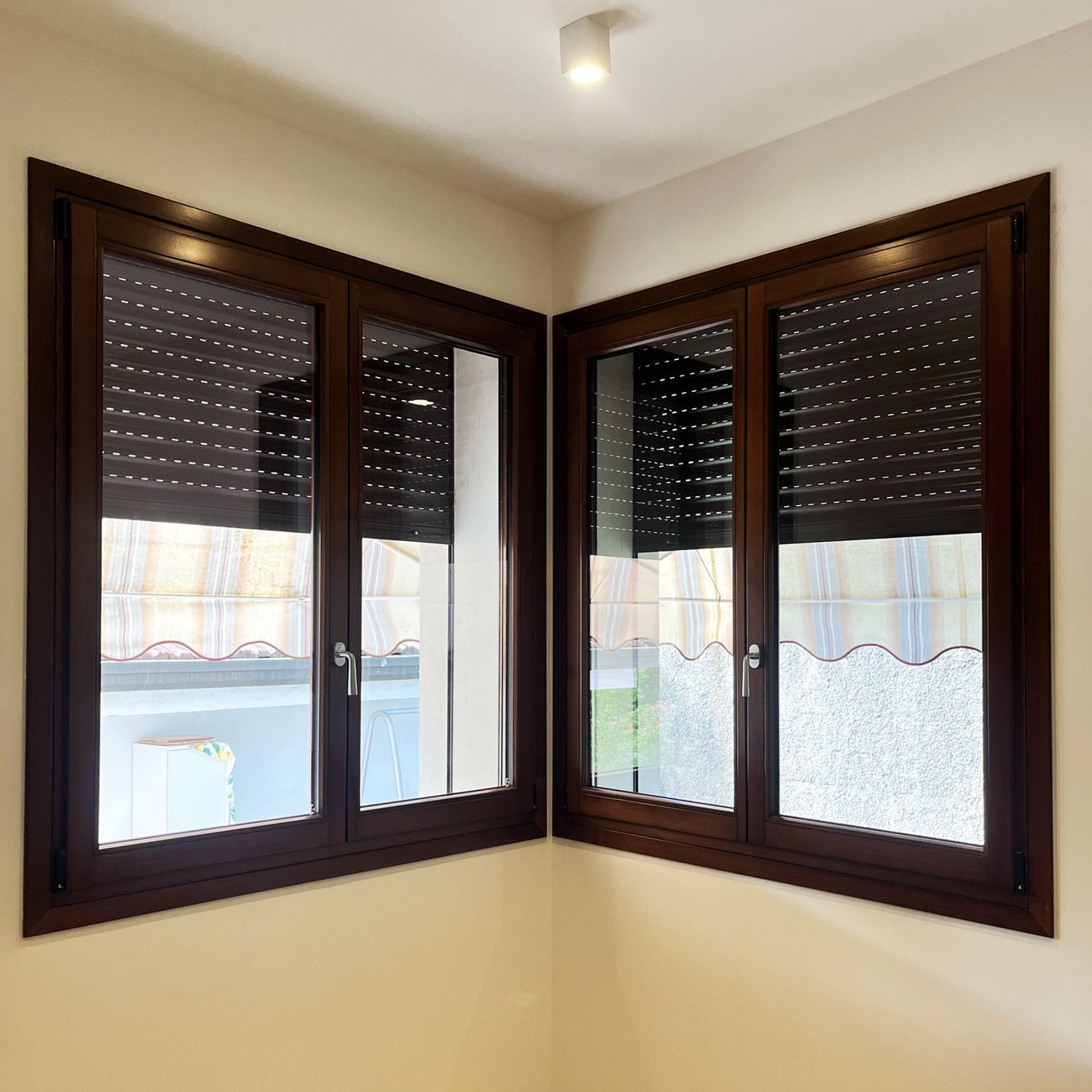 Due finestre con cornici marroni che si incontrano ad angolo, dotate di persiane avvolgibili a lamelle scure, parzialmente abbassate.