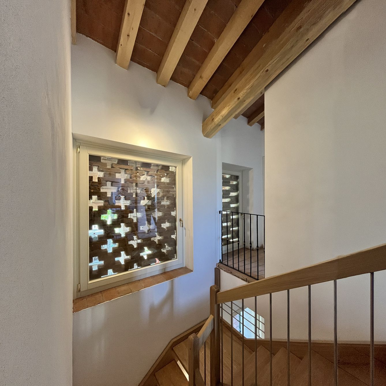 Una scala con travi in ​​legno e una parete bianca, caratterizzata da una finestra con un paravento decorativo a motivo incrociato che garantisce la privacy.