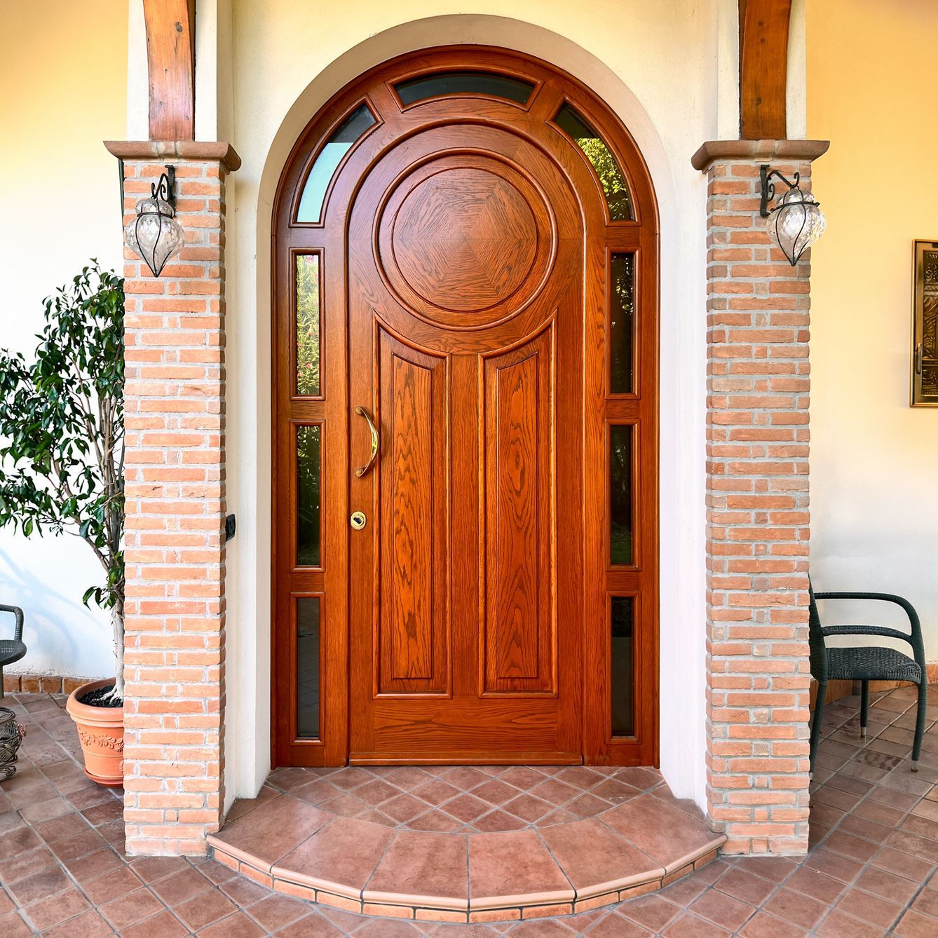 Una porta d'ingresso ad arco in legno marrone chiaro con pannelli in vetro e un disegno circolare, fiancheggiata da due pilastri in mattoni con lampade.