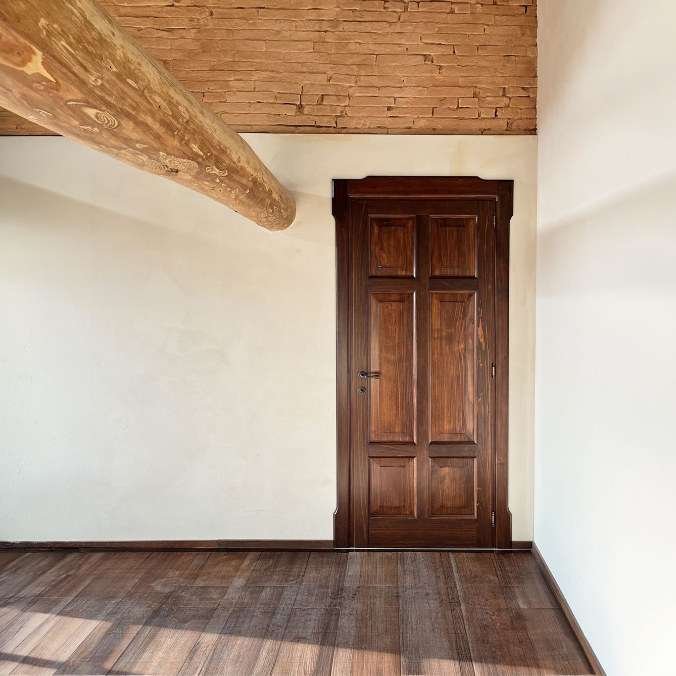 Una porta di legno scuro in una stanza con pareti bianche, pavimento in legno e una grande trave rustica sul soffitto.
