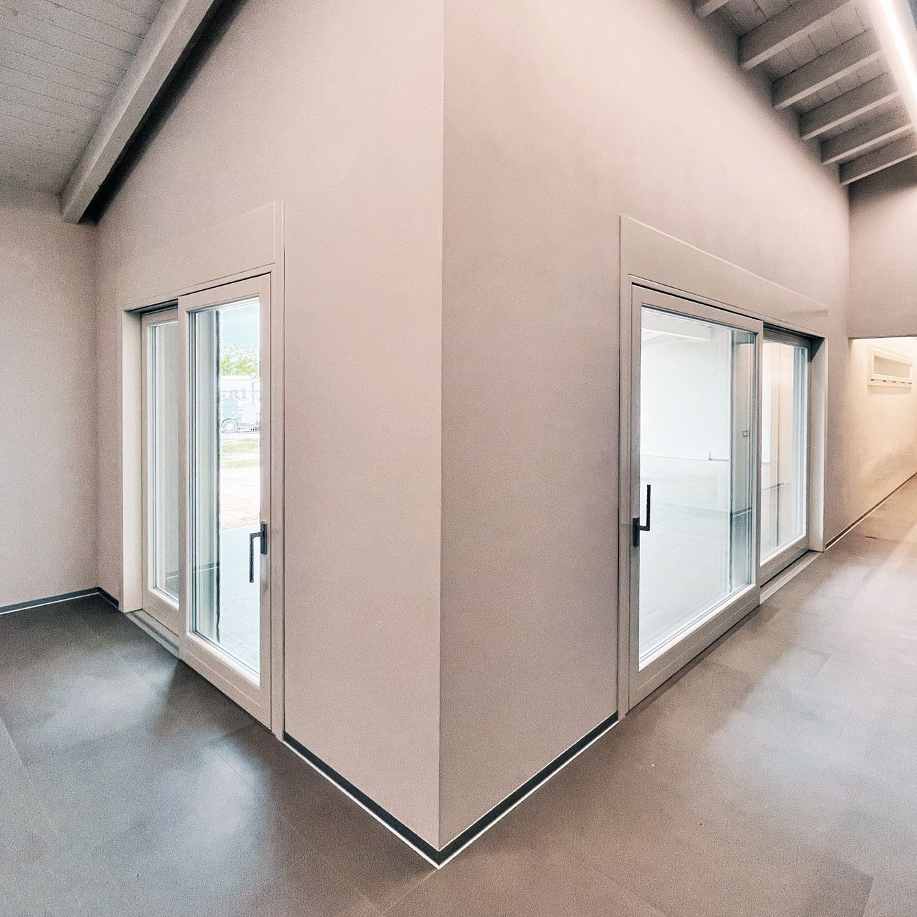 Un angolo di un corridoio moderno e minimalista con pareti beige lisce, pavimento in piastrelle grigie e porte a vetri con cornici sottili.