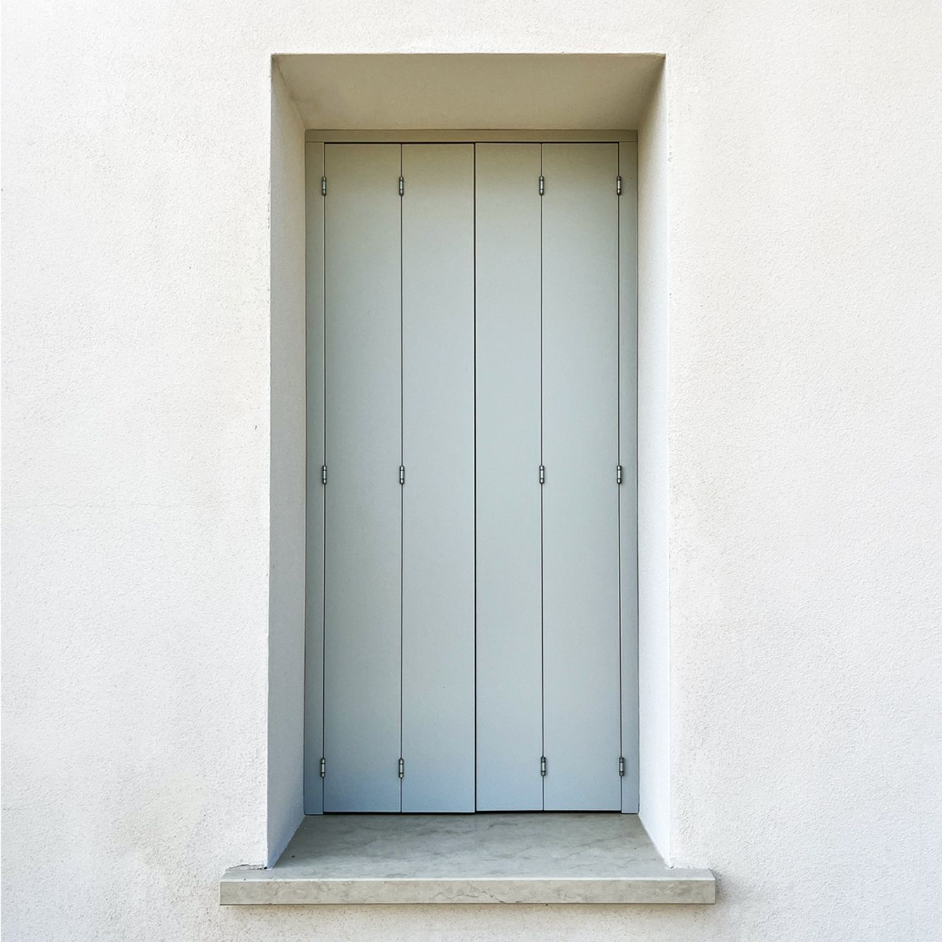 Persiane doppie chiuse di colore grigio chiaro, incastonate in una parete bianca con finitura a rilievo, sopra un davanzale in cemento.