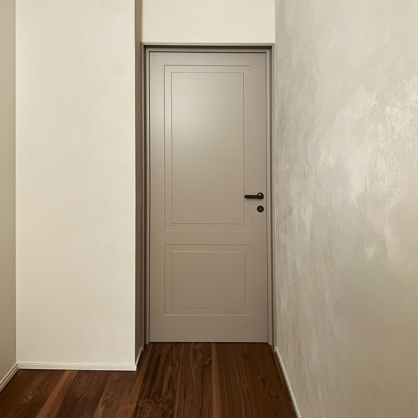 Una porta a pannelli grigio chiaro incastonata tra una parete bianca liscia e una parete con superficie ruvida, sopra un pavimento in legno scuro.