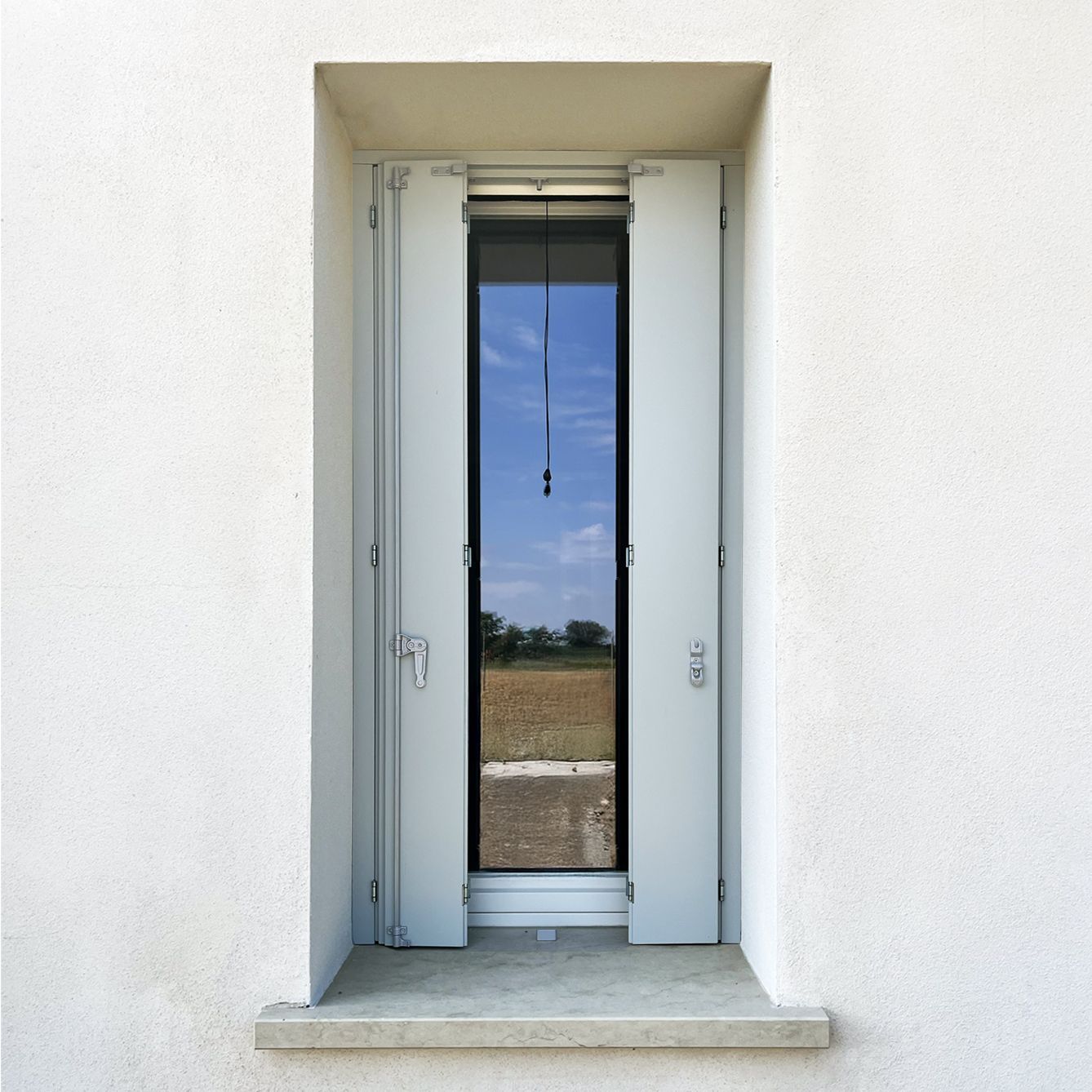 Un portone esterno alto e stretto, di colore grigio chiaro, con persiane aperte, incastonato in un muro di stucco bianco, che incornicia un paesaggio rurale.