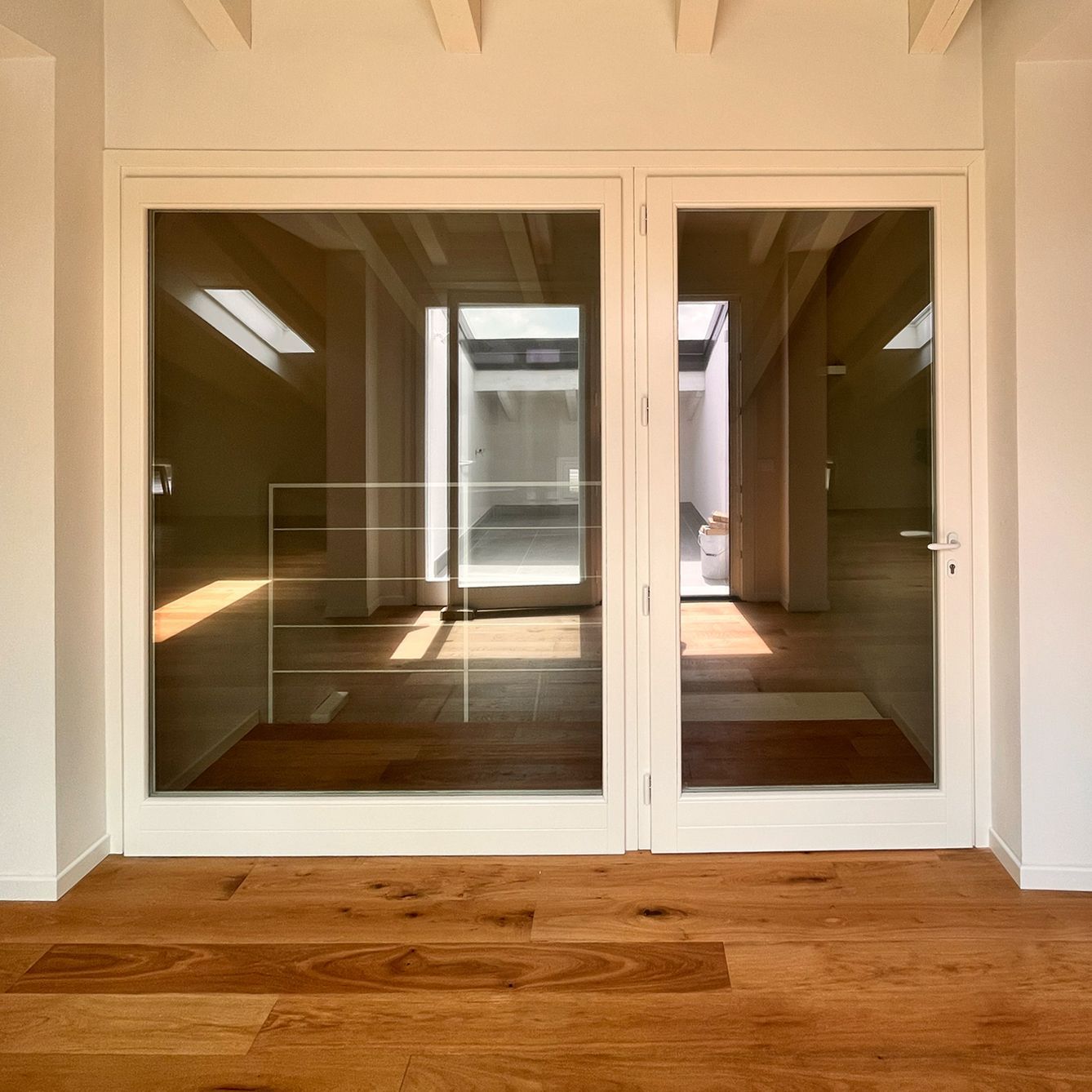 Una coppia di porte a vetri con cornice bianca, incastonate in una parete bianca, lasciano intravedere, attraverso i vetri, una stanza luminosa e vuota con pavimento in legno.