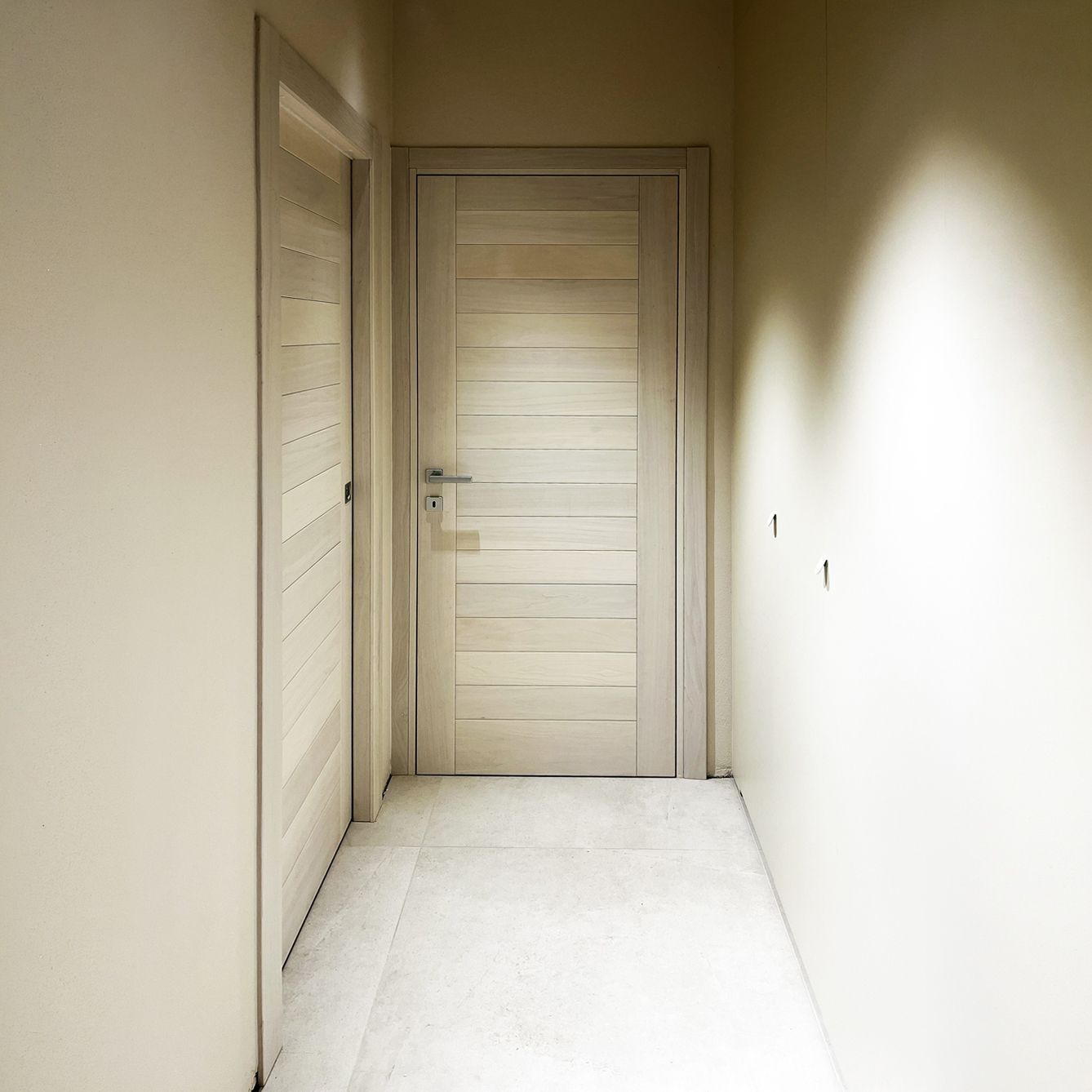 Un corridoio stretto con pareti e pavimento chiari, caratterizzato da due porte di legno con pannelli orizzontali.