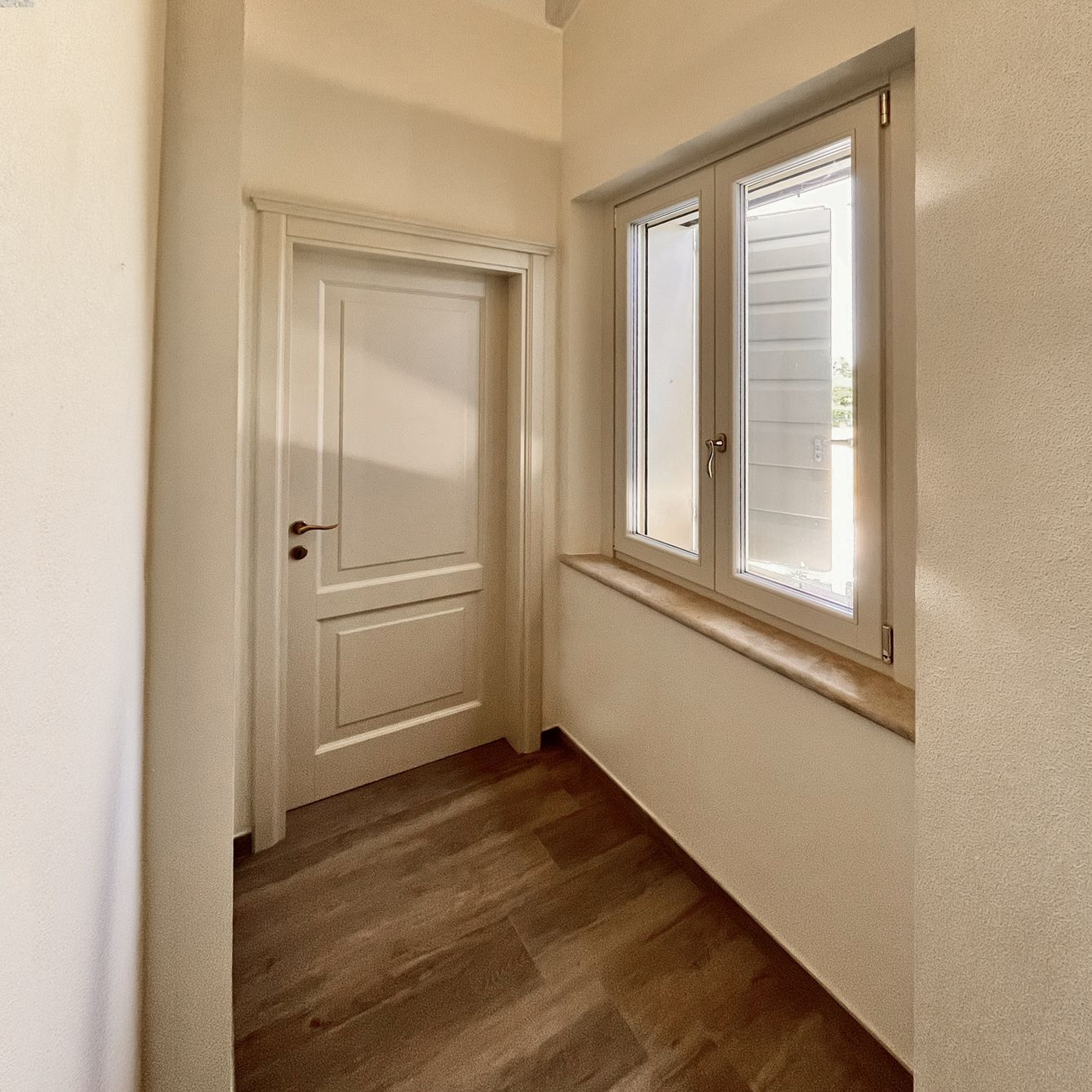 Un corridoio con pareti chiare e pavimento effetto legno, caratterizzato da una porta bianca chiusa e una finestra con persiane.