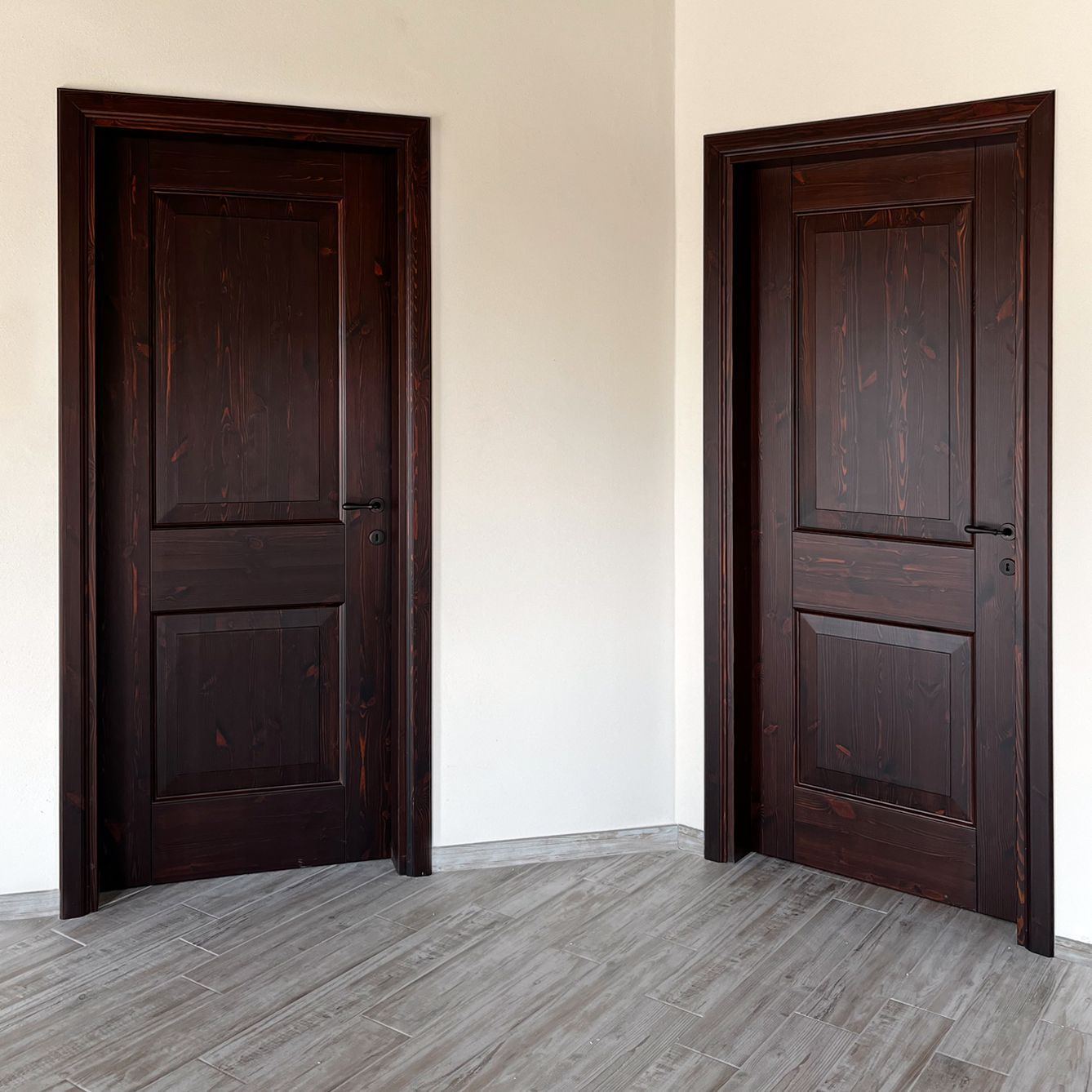Due porte in legno scuro con modanature decorative su una parete bianca sopra un pavimento grigio effetto legno.
