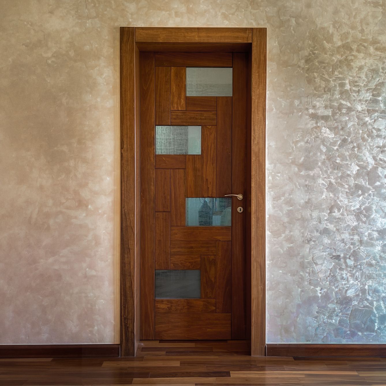 Una porta in legno scuro con sei pannelli rettangolari, inserita in una stanza rustica con pareti color crema e pavimento in legno.