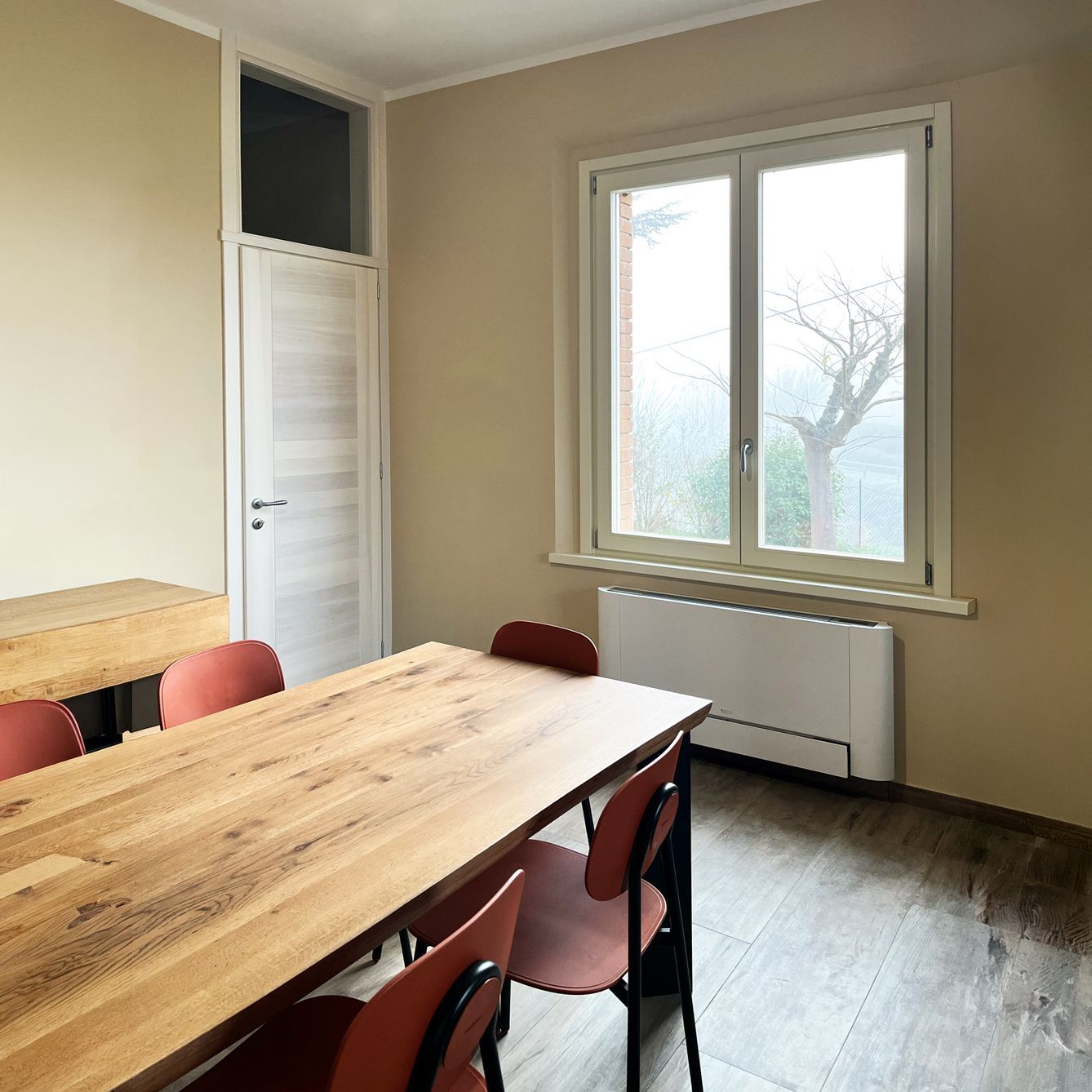 Una sala riunioni con un tavolo in legno chiaro, sedie color ruggine, una finestra con vista e un termosifone bianco su una parete beige.