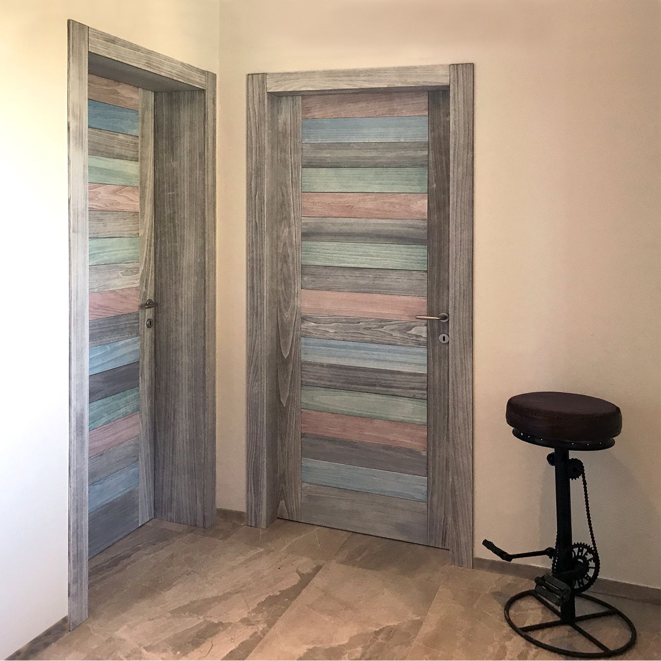 Due porte interne con assi di legno orizzontali multicolori in una cornice grigia, accanto a uno sgabello in stile industriale.