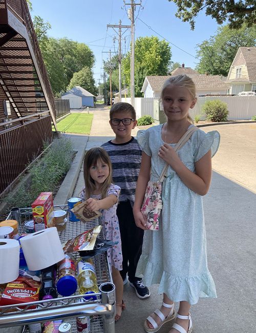 A boy and two girls are standing next to a shopping cart full of food from the blessing box
