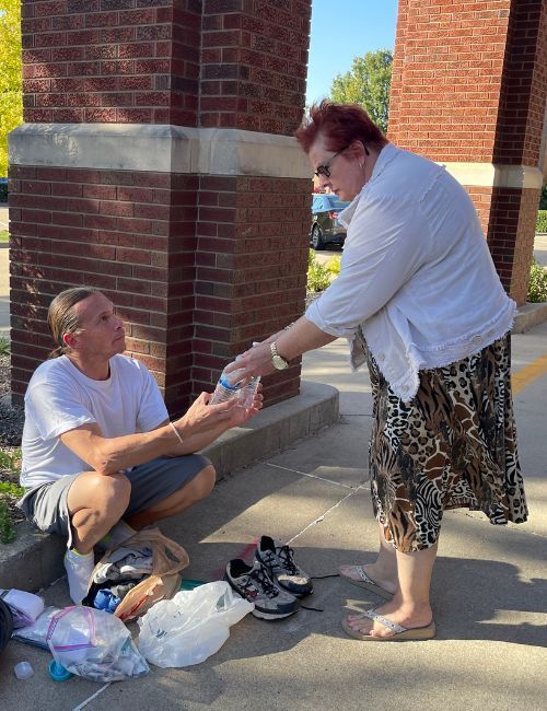 A woman is giving a bottle of water to a man sitting on the sidewalk.