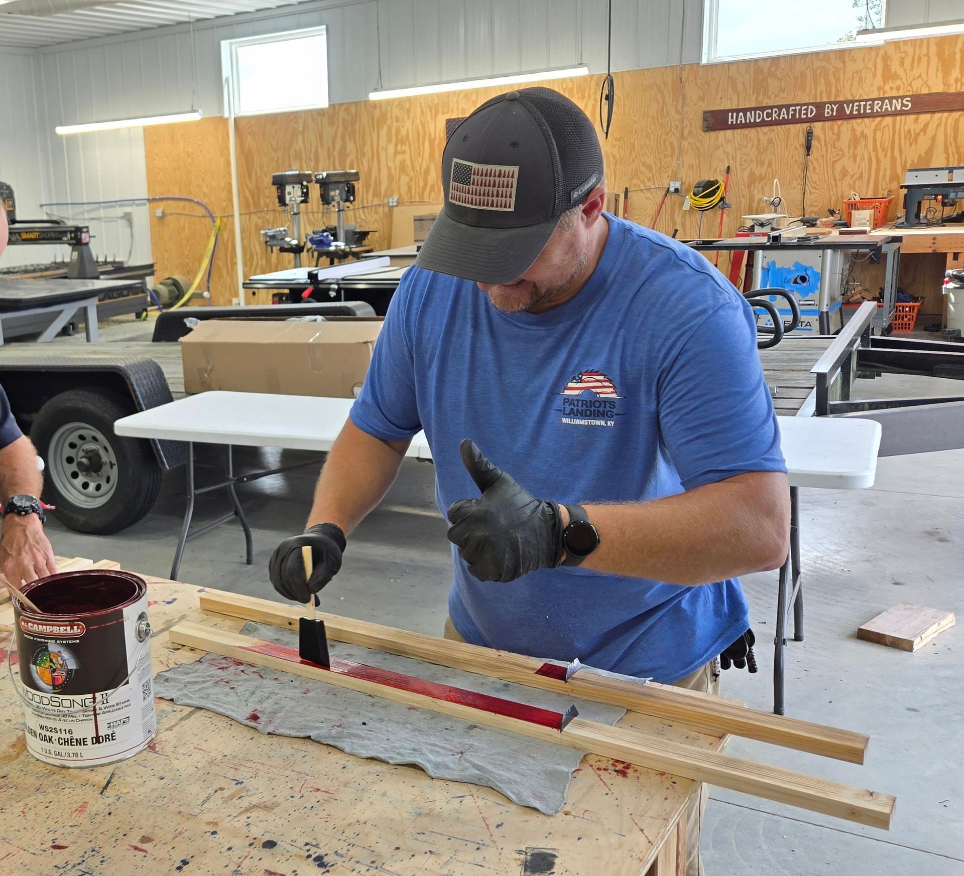 Man in blue shirt paints a wooden plank red inside a workshop, giving a thumbs-up.