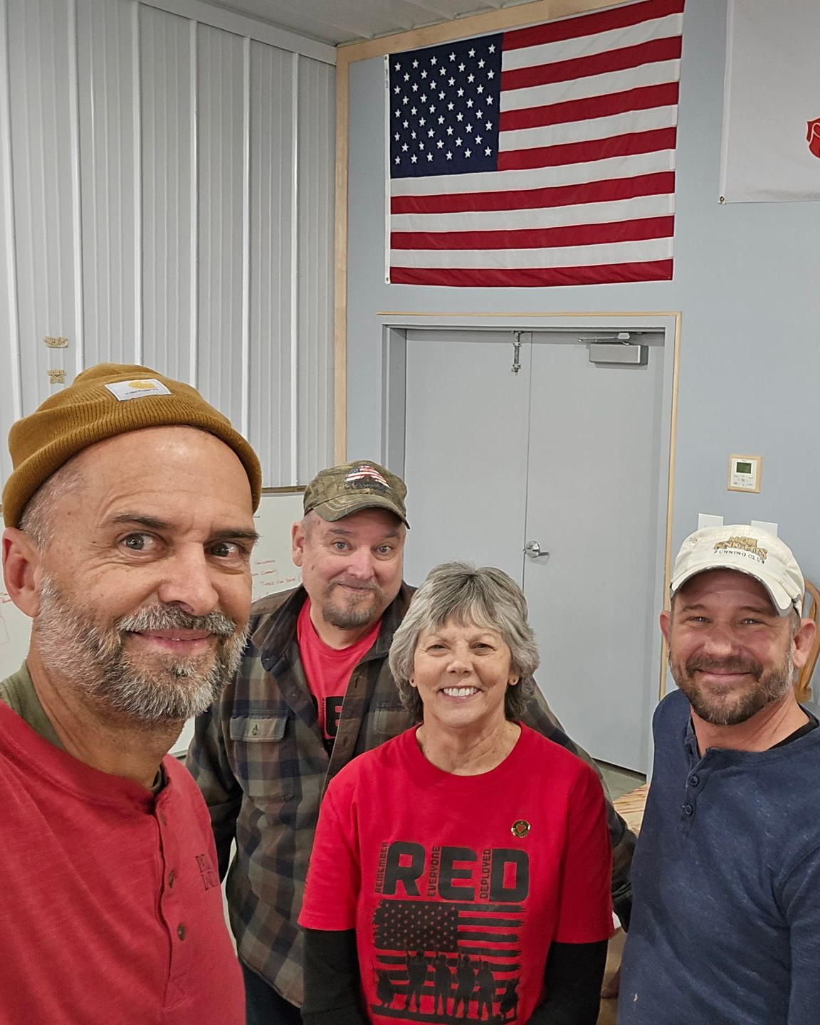 Four people smiling, indoors, near an American flag. One wears a red shirt that reads