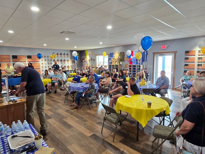 Large gathering inside, people seated at tables, blue and yellow decor.