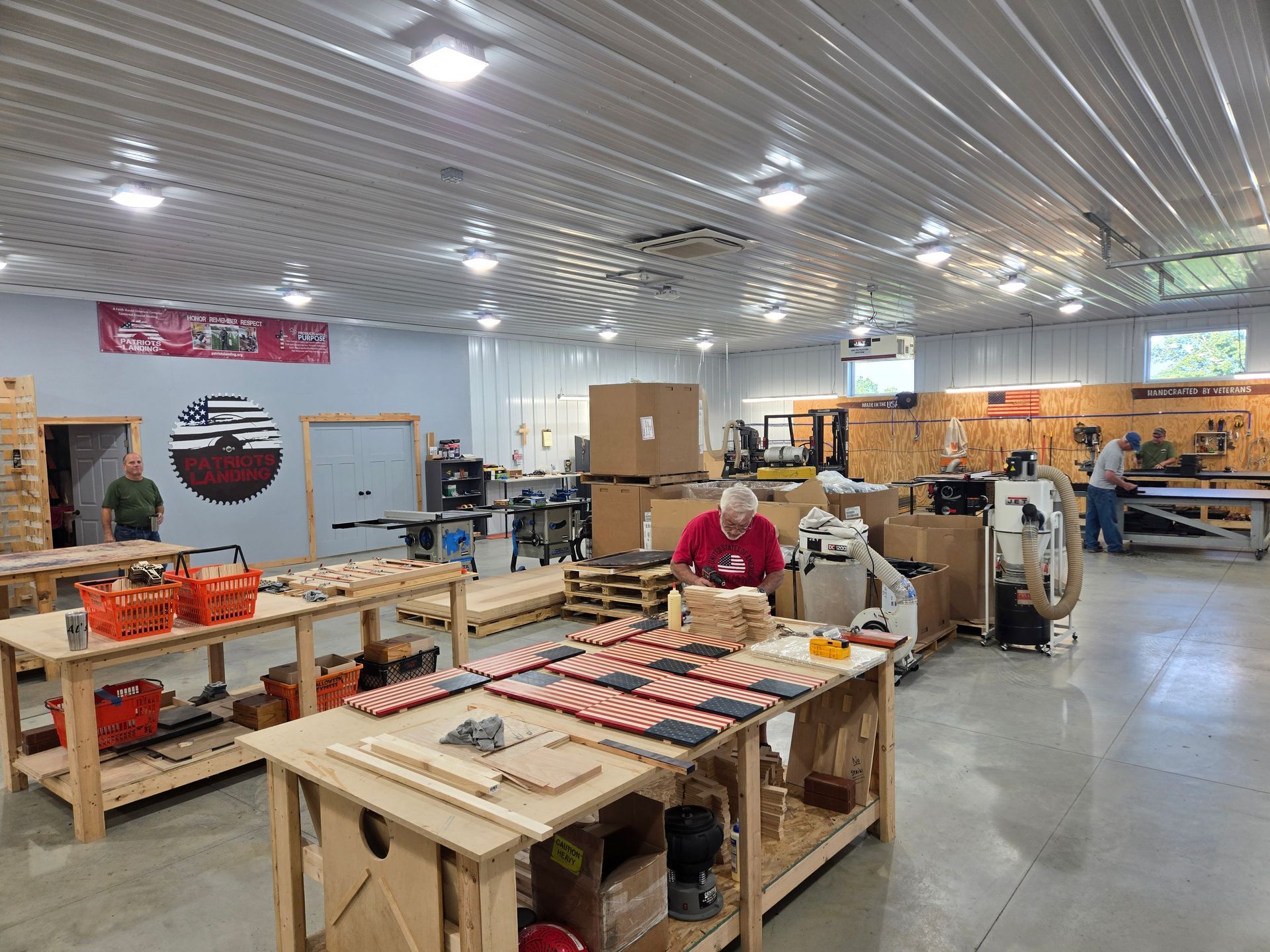 Workshop interior: people assembling wooden American flags at workbenches with tools, boxes, and supplies.