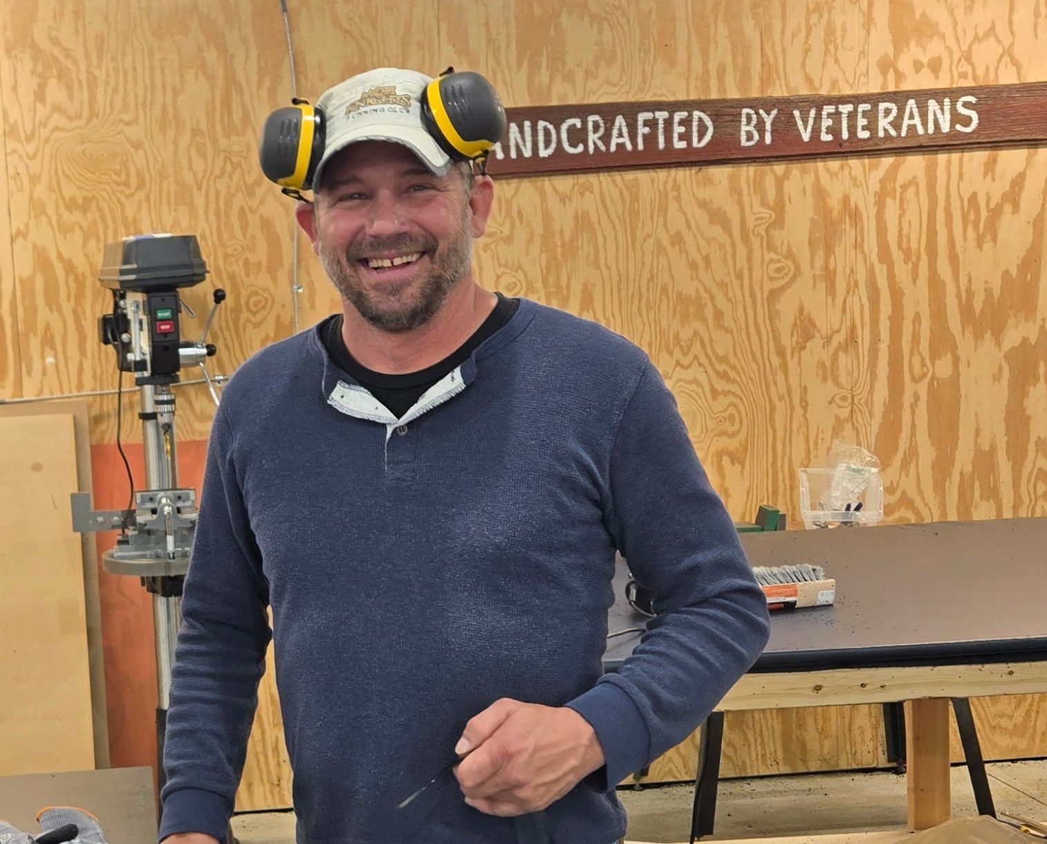 Man in workshop wearing earmuffs, smiling. Sign says