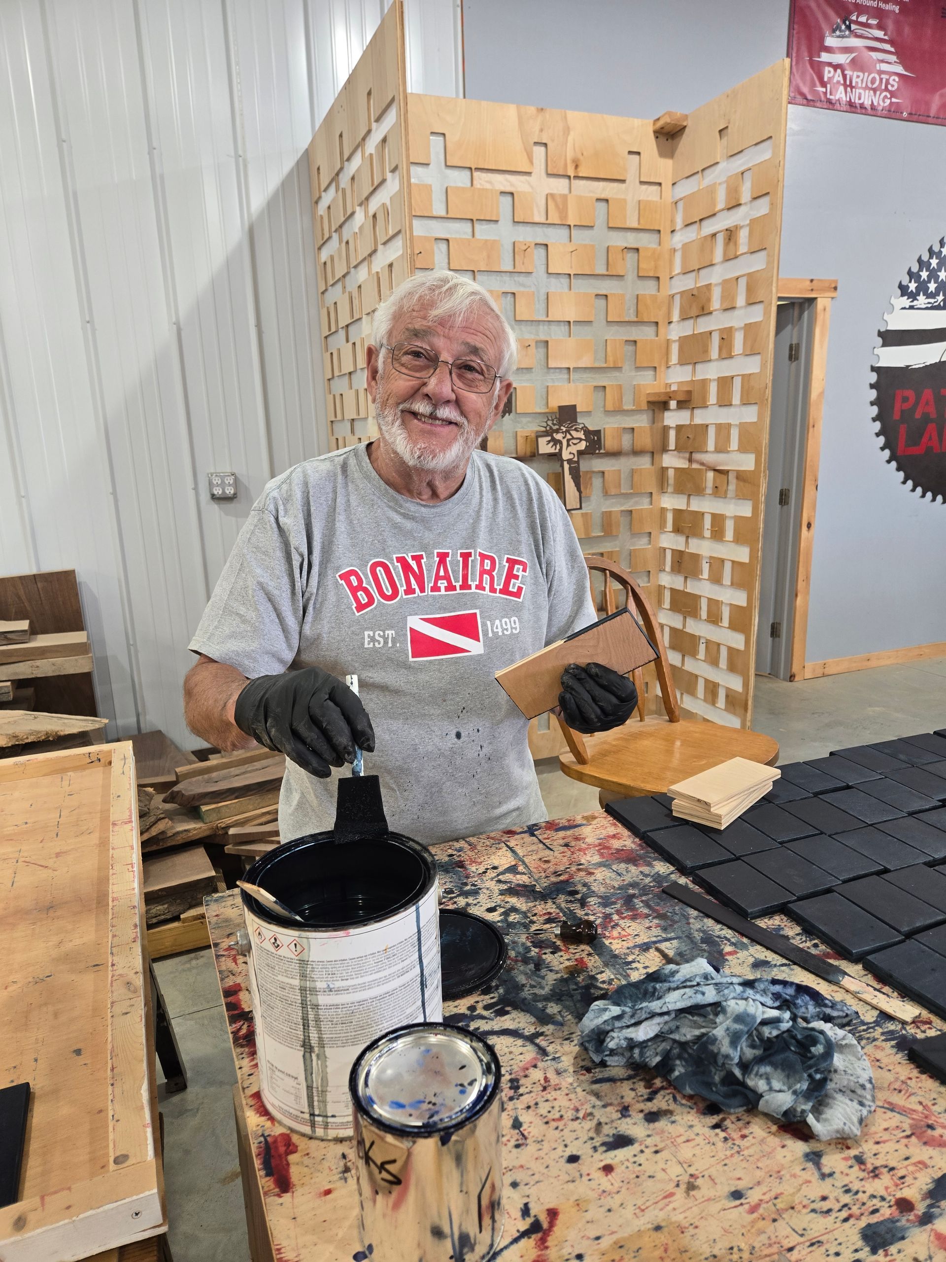 Smiling man painting wood squares with black paint, in a workshop. He's wearing a gray shirt and gloves.