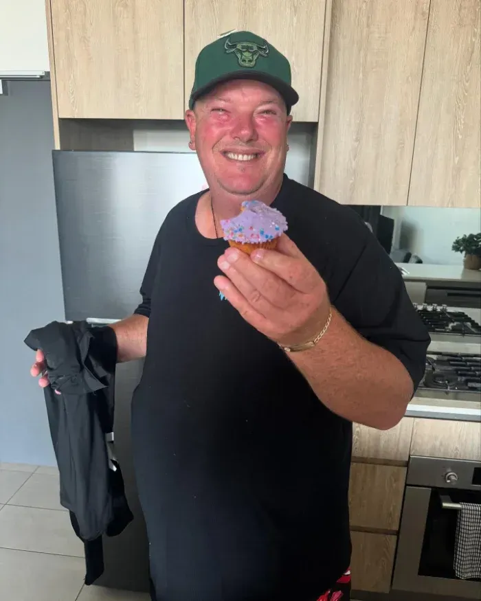 Man in black shirt and green cap holding a purple frosted cupcake, smiling indoors — Mentally Hooked in Brisbane, QLD