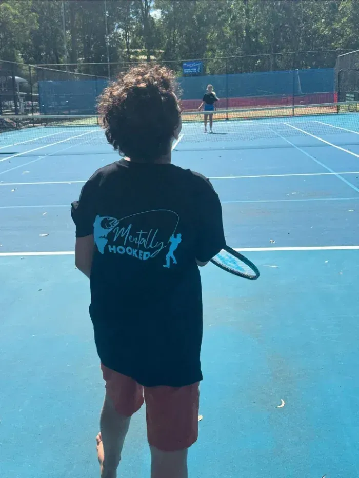 Boy on a blue tennis court holding a racket, facing another player in the distance — Mentally Hooked in Helensvale, QLD