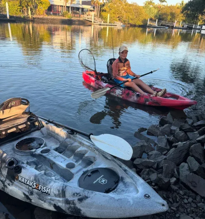 Kayaker on a red kayak with a net, next to a camo kayak on shore in a canal — Mentally Hooked in Brisbane, QLD