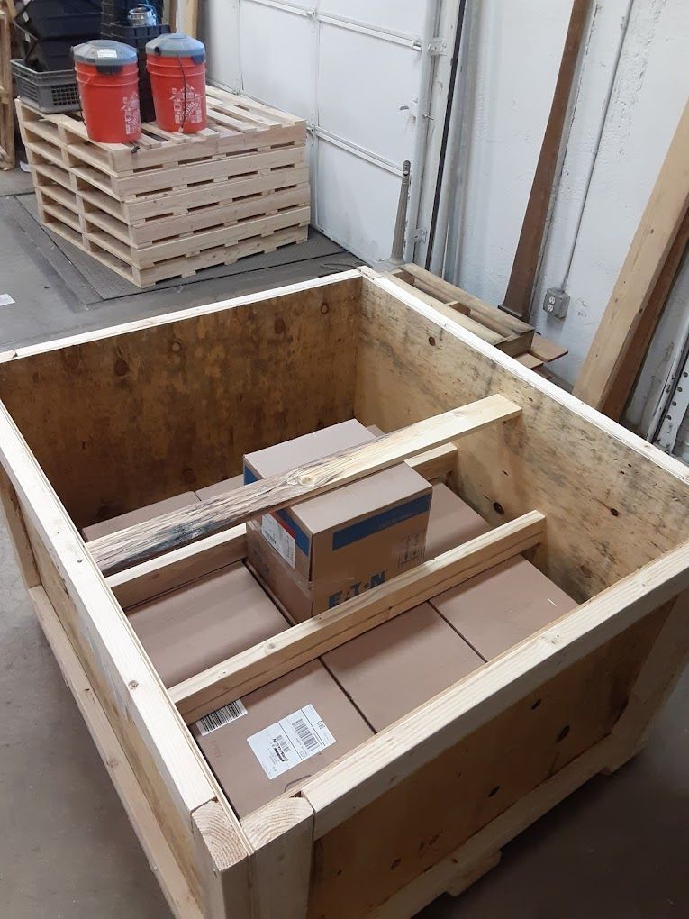Wooden crate containing cardboard boxes, with additional boxes and buckets on a pallet in the background.