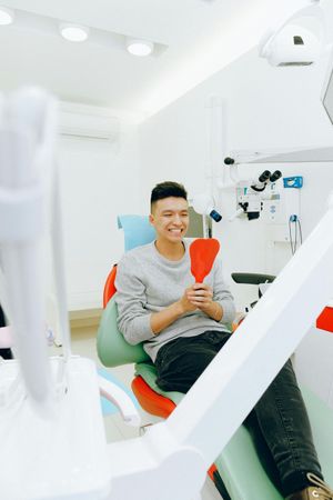 Man in dental chair, holding a red mirror, smiling. Dental office setting.