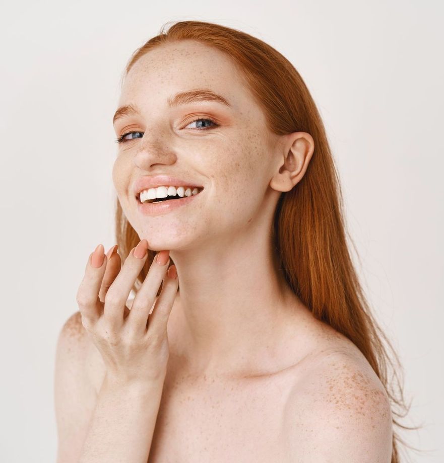 A woman with red hair and freckles is smiling and touching her face.