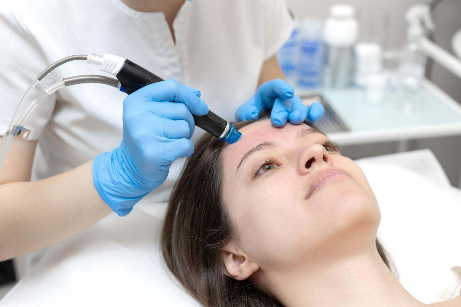 Person receiving a facial treatment with a handheld device. Blue-gloved hands, light skin, white setting.