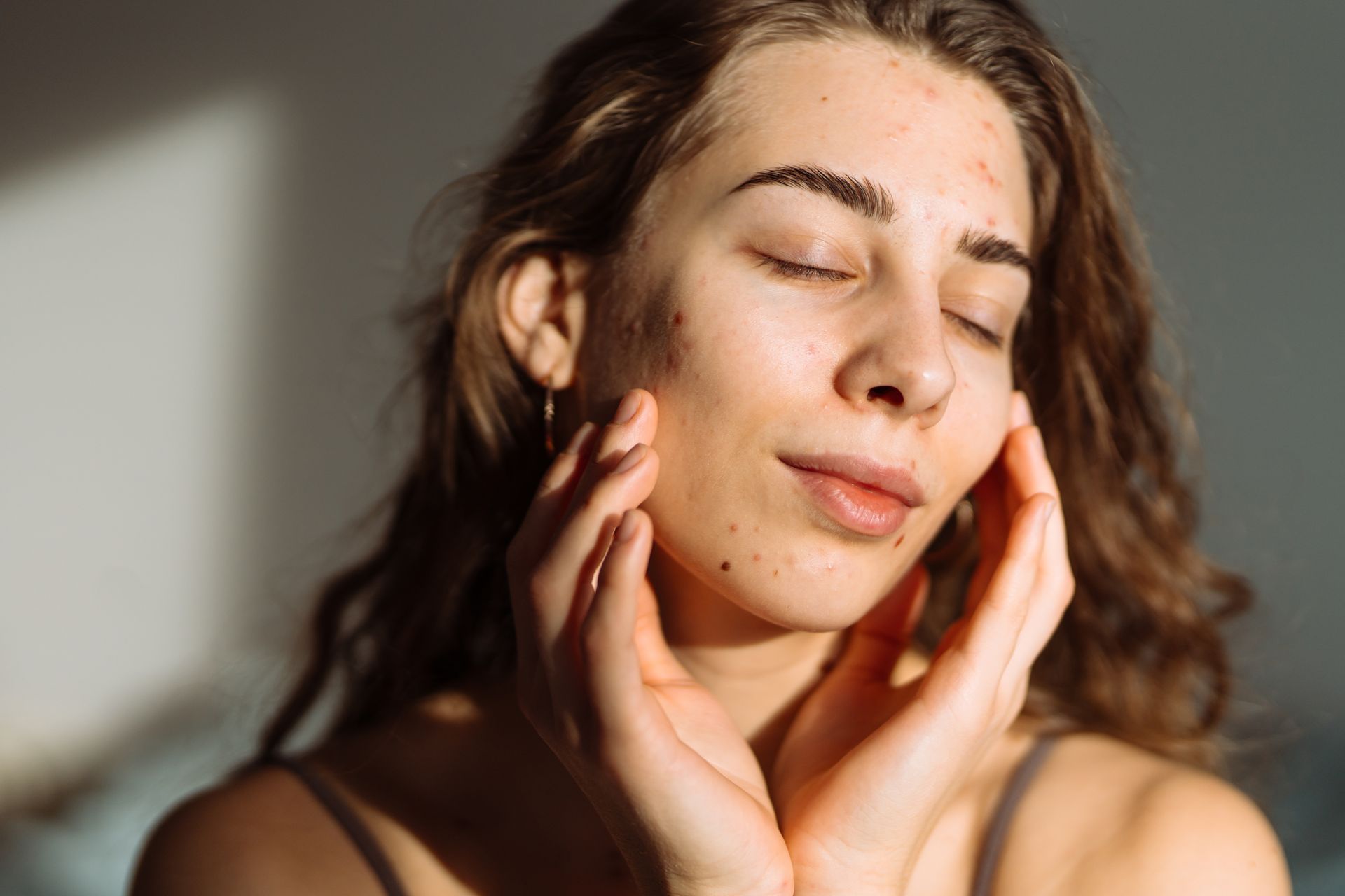 Woman with eyes closed gently touching her cheeks in warm sunlight.