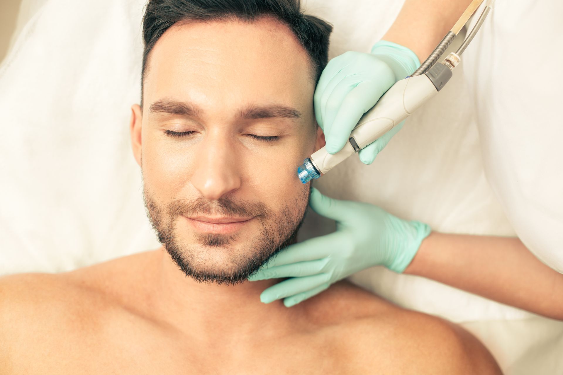 Man receiving facial treatment; spa setting. Face close-up with closed eyes, being treated with blue-handled tool.