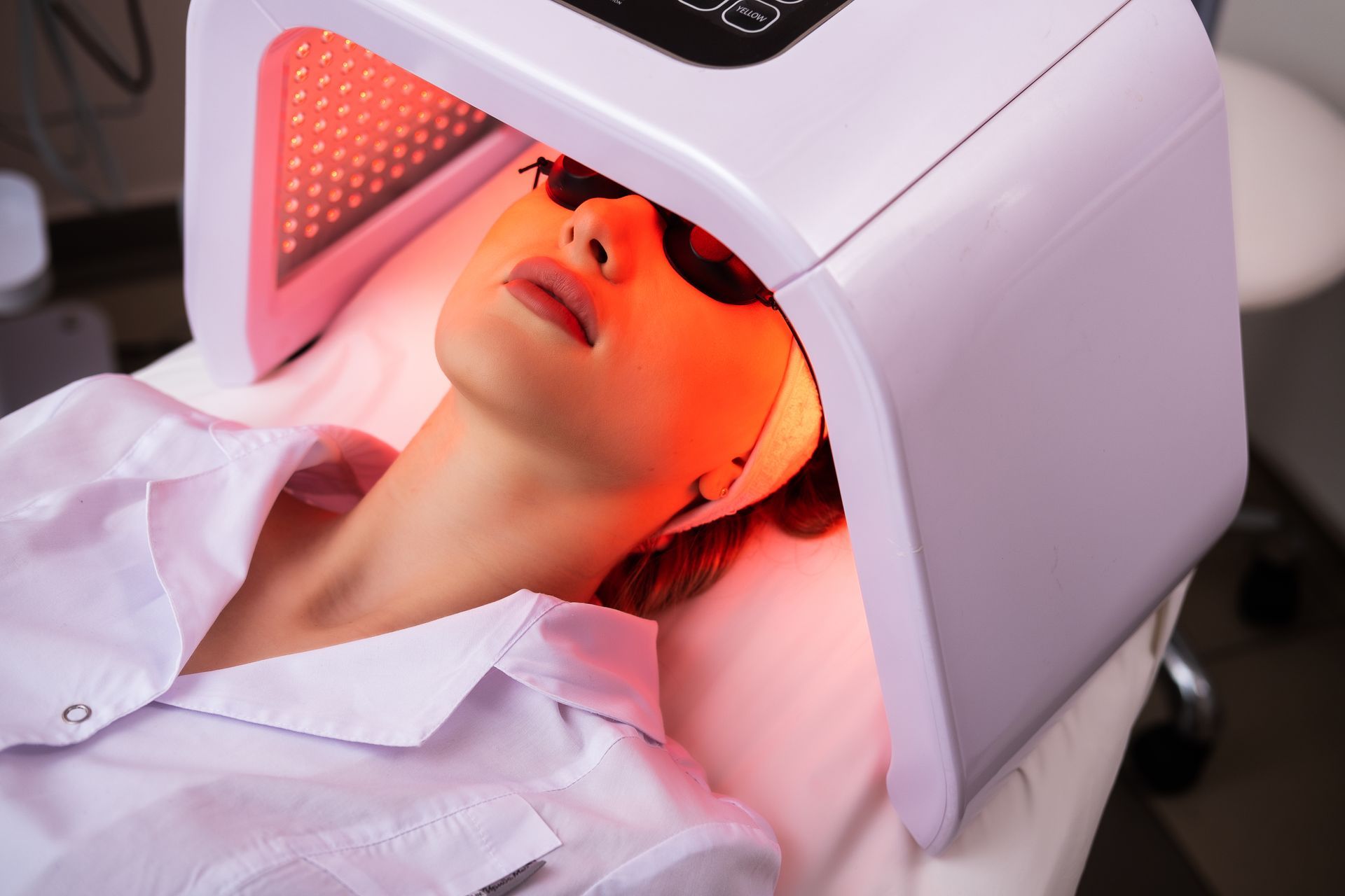 Woman under red LED light therapy device, wearing eye protection, in a spa setting.