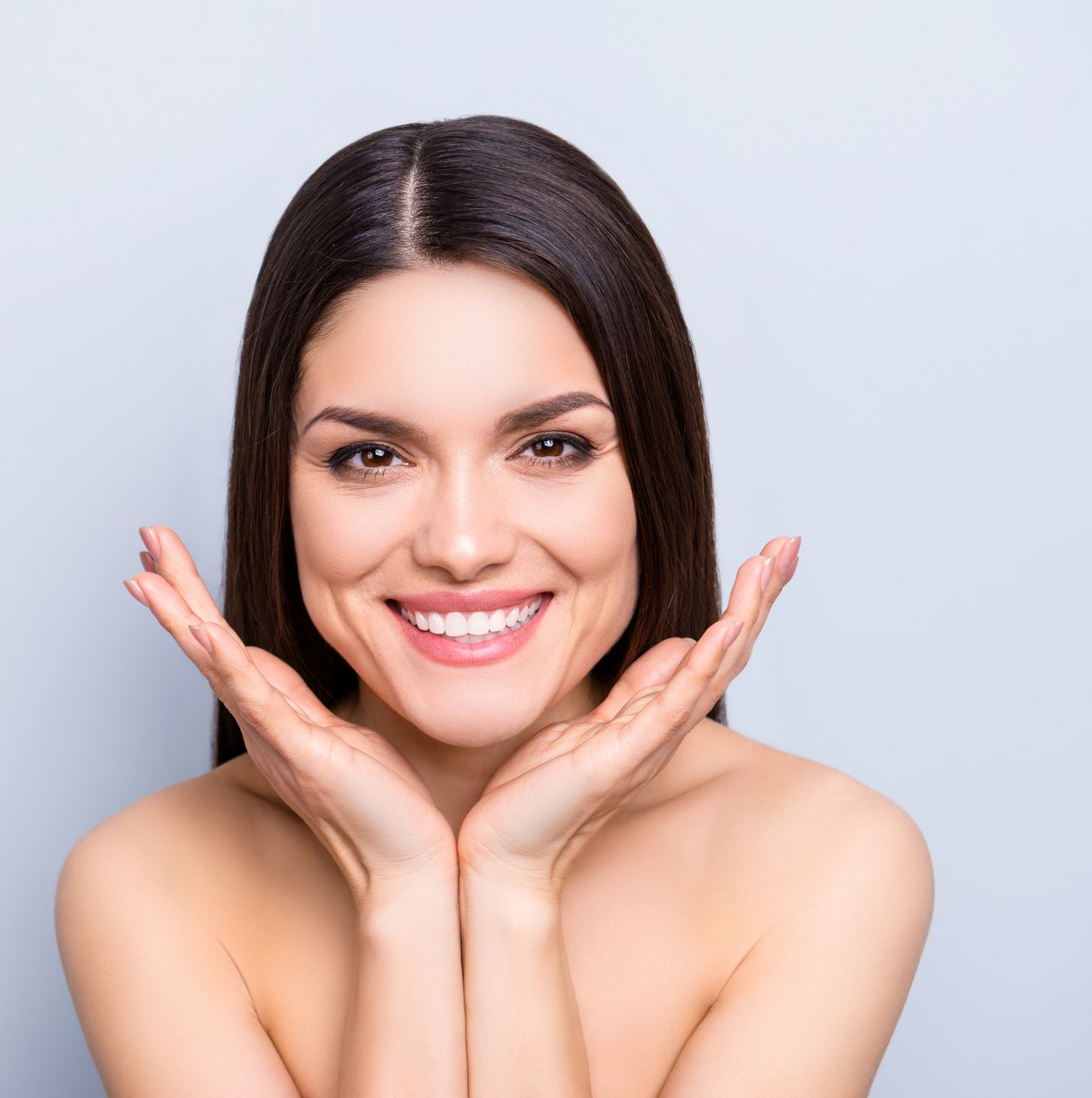Woman with dark hair smiles, holding hands near face; bare shoulders against a light gray background.