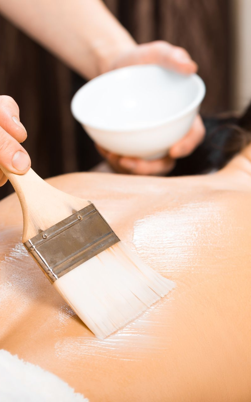 A person receiving a spa treatment. A brush applies white substance to the back. A bowl is held nearby.