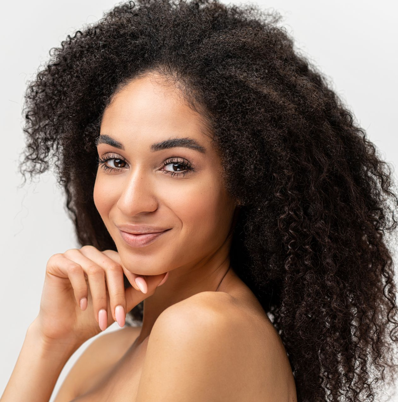 Person with curly hair smiles, hand to chin, against a white background.