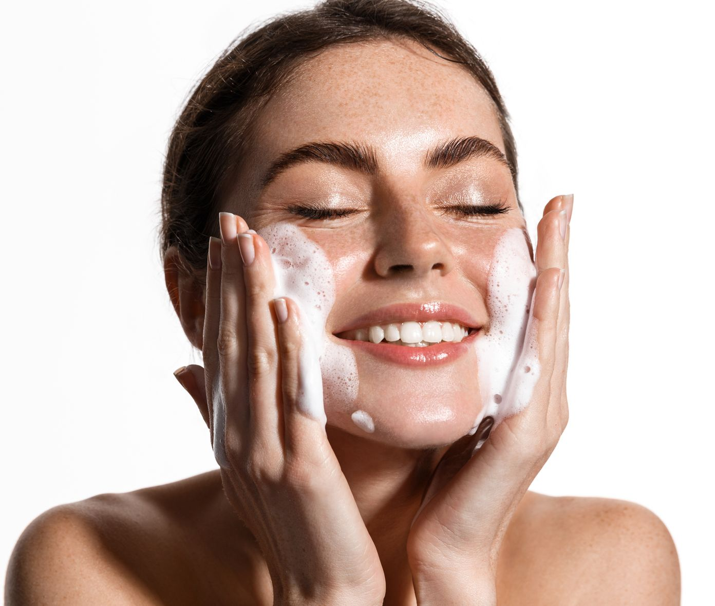 Woman with eyes closed, smiling, washing face with foamy cleanser. White background.