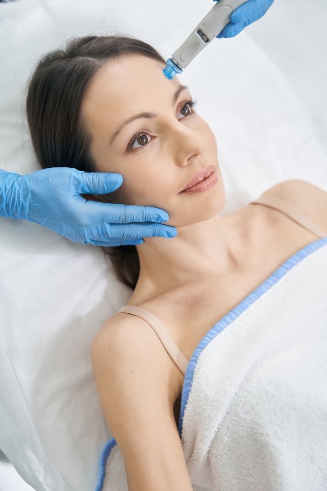 Woman receiving facial treatment; blue gloved hands, skincare device, white towel, lying down.