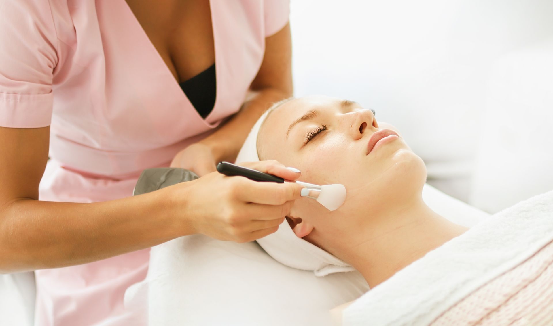 Woman receiving a facial treatment in a spa; esthetician uses a brush.