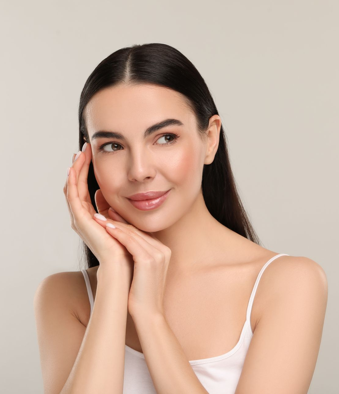 Woman with dark hair touches her face, looking to the side, wearing a white tank top.