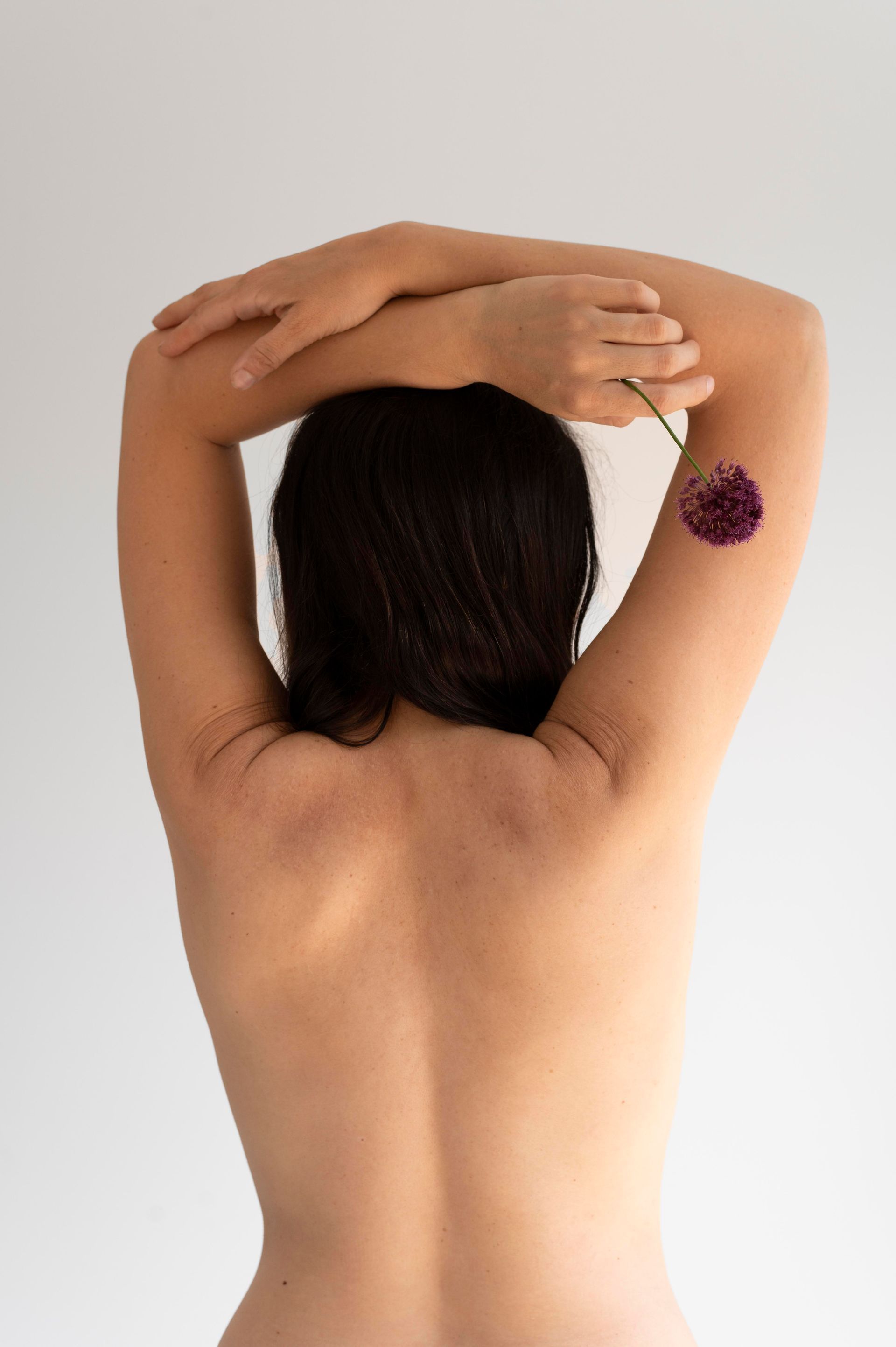 Naked torso of a person, back to the camera, arms crossed overhead, holding a purple flower.