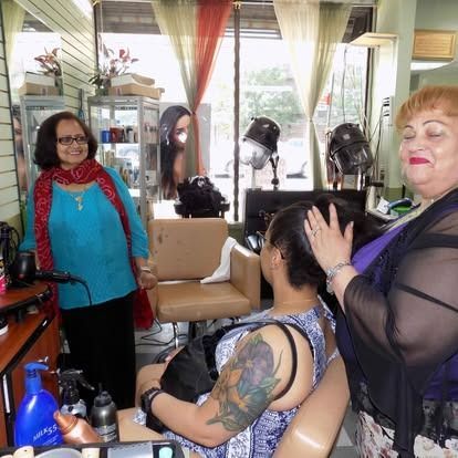 Inside a hair salon, two women are styling a customer's hair. A woman in blue watches, smiling.