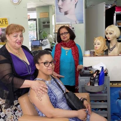 Three women in a salon. A woman in a blue dress is having her hair styled. Two other women are standing.