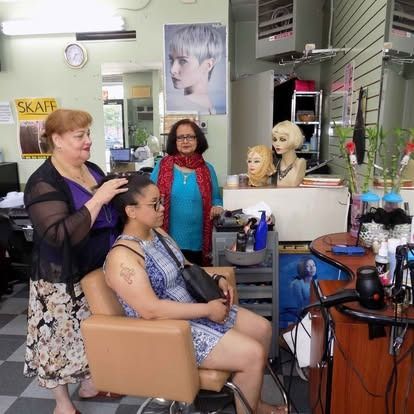 Salon scene: Three women, two styling a client's hair. Woman in blue dress sits, two women stand; one checks hair.