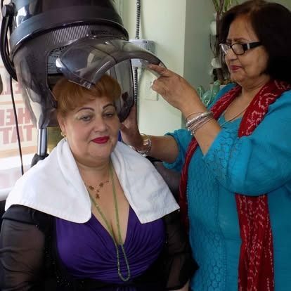 Woman under salon hair dryer, another adjusting it. Both women are smiling indoors.