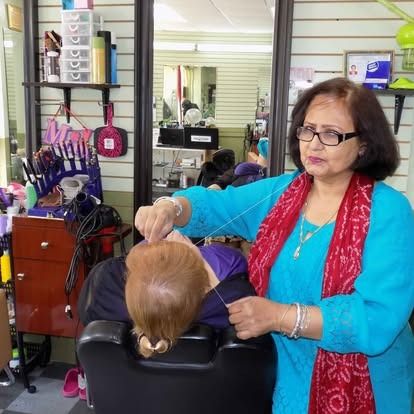Woman threading eyebrow of a mannequin in a salon. She wears blue, red scarf, glasses.