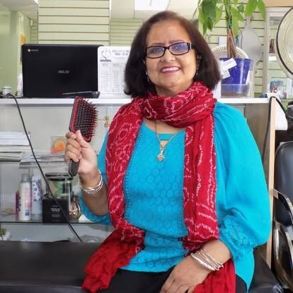 Woman with glasses smiling, holding hairbrush, wearing blue top and red scarf in a salon.